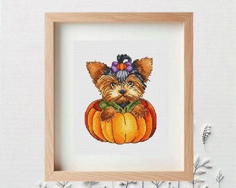 May include: Cross-stitch picture of a Yorkshire Terrier puppy in an orange pumpkin. The puppy has brown and black fur, wearing a purple and orange flower headband. The artwork is framed in a light brown wooden frame.