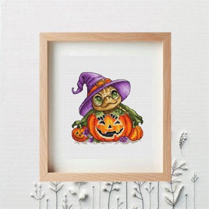 Puede incluir: Arte de punto de cruz de una tortuga con un sombrero de bruja morado, asomándose por detrás de una calabaza naranja tallada. Una calabaza más pequeña está a la izquierda. La obra está enmarcada en un marco de madera marrón claro.