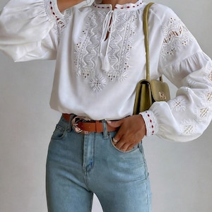 May include: A white long-sleeve blouse featuring intricate white embroidery, cut-out details on the sleeves, and a tie-neck closure. The blouse is paired with light blue denim jeans, a brown leather belt with a gold buckle, and a green shoulder bag.