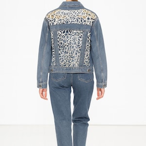 Op de afbeelding: Een blauwe spijkerjas met een decoratief wit kanten achterpaneel, gecombineerd met bijpassende blauwe jeans. De jas heeft lange mouwen en een klassieke kraag. Het model draagt witte sneakers. De outfit is casual en stijlvol.