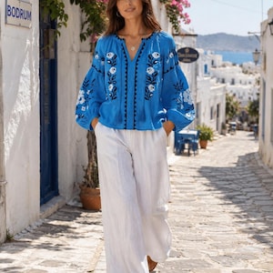 May include: A bright blue long-sleeved blouse featuring a deep V-neckline with dark blue trim and white floral embroidery on the front and sleeves, paired with loose-fitting white wide-leg trousers that reach the floor.