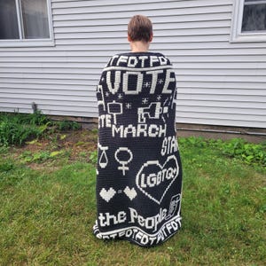 Puede incluir: Una manta de ganchillo en blanco y negro con la palabra "VOTE" y otros símbolos de protesta. La manta también presenta las palabras "MARCH", "LGBTQ" y "the People".