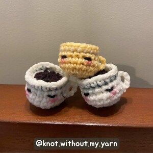 May include: Three crocheted coffee cups in white, yellow, and white with dark brown filling. Each cup has a smiling face with pink cheeks and black eyes. The cups are stacked on top of each other. The text "@knot.without.my.yarn" is at the bottom.