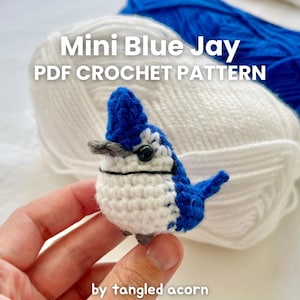 以下が含まれることがあります： 手で持たれた、白と青のディテールが施されたかぎ針編みの小さな青い鳥。鳥は黒い目と灰色のくちばしをしています。「Mini Blue Jay PDF Crochet Pattern」の文字が鳥の上にあり、「by tangled acorn」が下にあります。
