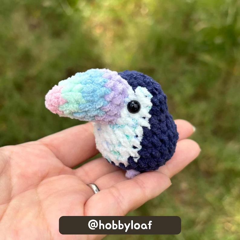 Cool-toned crochet toucan plush held in a hand against a grassy background, with the tester’s Instagram name shown at the bottom.