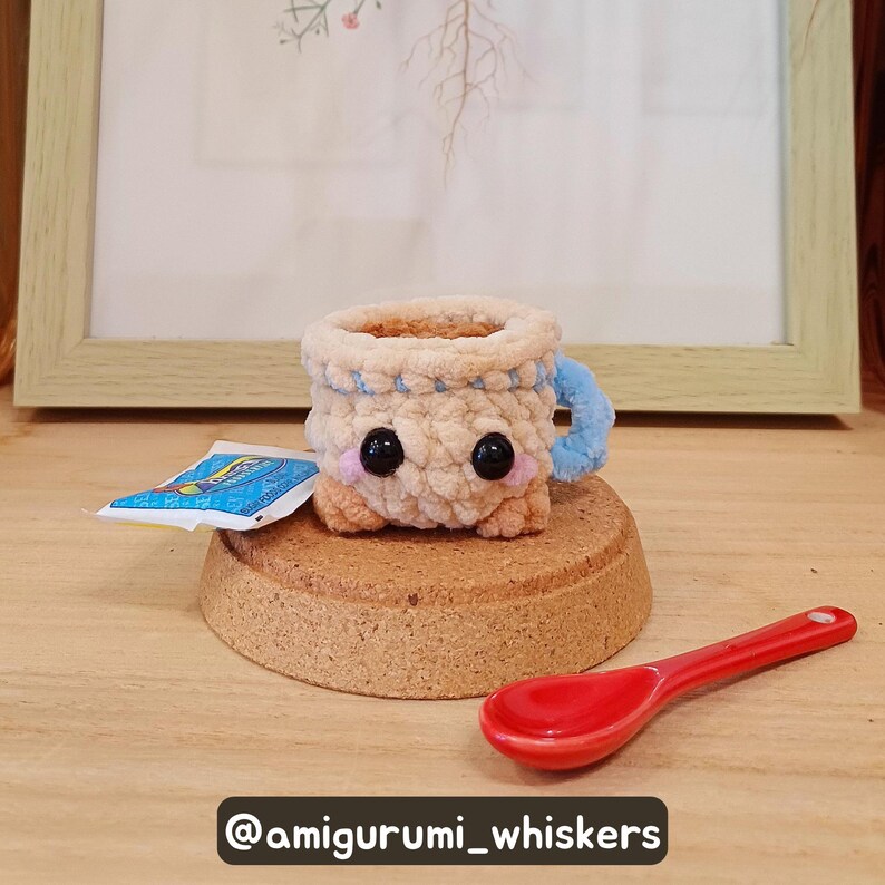 May include: A crocheted coffee cup amigurumi with black eyes and pink cheeks, sitting on a cork coaster. A small blue and white packet and a red spoon are next to the cup. The cup has a blue handle and trim. The text "@amigurumi_whiskers" is at the bottom.
