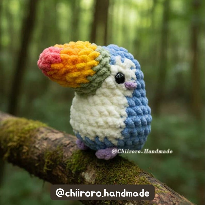 Pastel-colored crochet toucan with a soft blue body and rainbow beak sitting on a branch against a forest background, with tester credit text at the bottom.