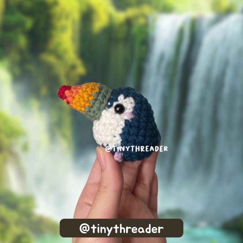Small chubby crochet toucan held between fingertips in front of a waterfall, showing the side profile with tester credit at the bottom.
