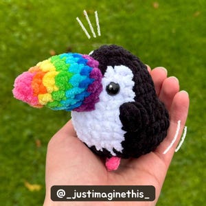 Crochet toucan with a saturated rainbow beak and pink feet held in a hand over grass, accented with white doodle details and tester name below.
