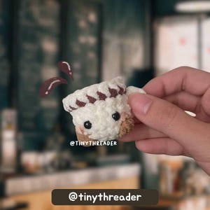 May include: A crocheted coffee cup with a chef's hat design. The cup is white with brown accents and black eyes. The text "@tinythreader" is visible. Two brown coffee splashes are floating above the cup.