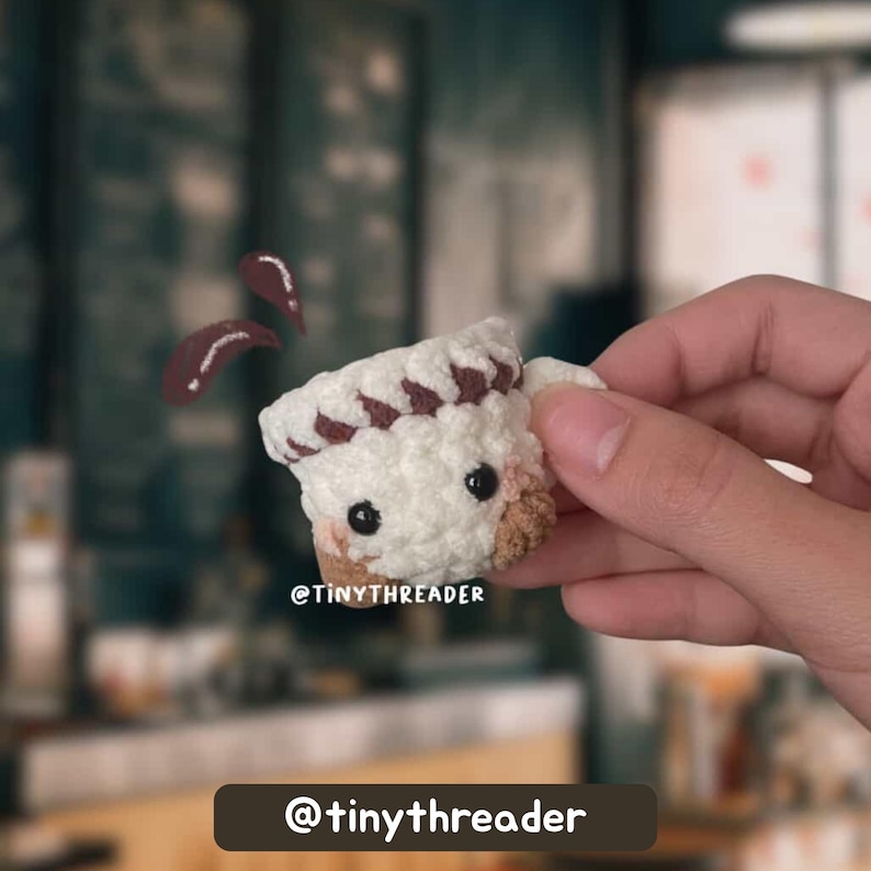 May include: A crocheted coffee cup with a chef's hat design. The cup is white with brown accents and black eyes. The text "@tinythreader" is visible. Two brown coffee splashes are floating above the cup.