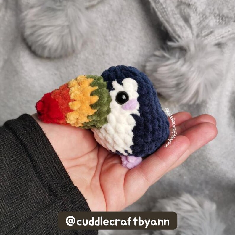 Rainbow toucan crochet plush in original pattern colors held in a hand against a soft, fluffy white background, with tester name displayed.