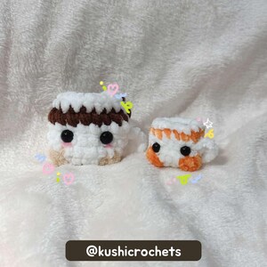 May include: Two crocheted, cube-shaped items resembling miniature desserts. One is white with a brown top and tan base, the other white with orange accents. Both have black eyes and are on a soft, white surface. The text "@kushicrochets" is at the bottom.