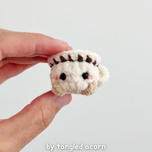 May include: A small, crocheted coffee cup with a cute face. The cup is primarily cream-colored with brown accents on the rim and base. It has black eyes and pink cheeks, giving it a friendly appearance. The text "by tangled acorn" is at the bottom.
