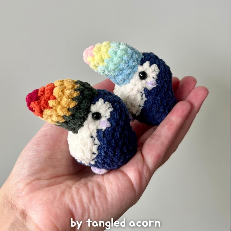 Two crochet toucans held in a hand — one with a bright rainbow beak and one with a pastel rainbow beak — shown from the side on a white background.