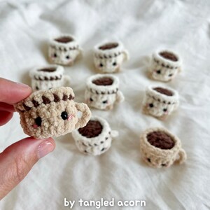 May include: A collection of small, crocheted coffee cups with smiling faces. The cups are beige with brown coffee and details. The text "by tangled acorn" is at the bottom of the image.