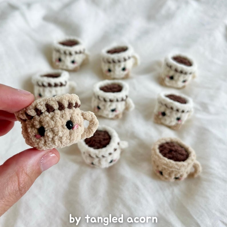 May include: A collection of small, crocheted coffee cups with smiling faces. The cups are beige with brown coffee and details. The text "by tangled acorn" is at the bottom of the image.