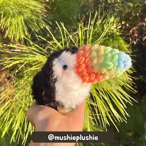 Crochet toucan with a black and white fluffy yarn body and rainbow beak held in front of a tree, with tester credit text at the bottom.