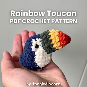 May include: A crocheted toucan, held in a hand, with a blue body, white chest, and a multi-coloured beak. The beak features yellow, orange, and red. The text "Rainbow Toucan PDF Crochet Pattern" is visible at the top.