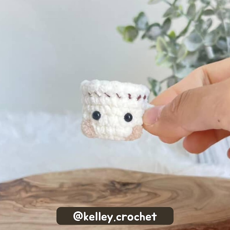 May include: A small, handmade crochet item in the shape of a cup or mug. The item is primarily white with brown accents and black eyes, and is held by a hand. The text "@kelley.crochet" is at the bottom.