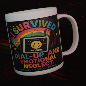 May include: White ceramic mug with a colorful design. The mug features the text "I SURVIVED DIAL-UP AND EMOTIONAL NEGLECT" with a rainbow, modem graphic, and the words "EMOTIONAL STATUS: buffering..."