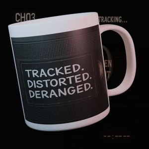 May include: White ceramic mug with a black textured design and the words "TRACKED. DISTORTED. DERANGED." printed in white. The mug has a white handle and rim. The word "tracking" is also visible.