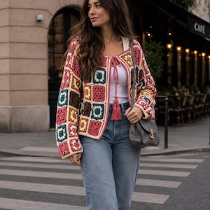May include: A long-sleeved crochet cardigan featuring a multi-coloured granny square pattern in shades of pink, teal, brown, and cream. The garment includes a front tie closure with pink tassels, worn over a white top and blue denim jeans.