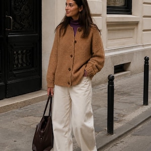 May include: A tan knit cardigan with a round neckline and brown buttons, worn over a purple mock neck top. The outfit includes wide-leg off-white trousers, brown trainers, and a dark brown leather tote bag carried by the side of the garment.