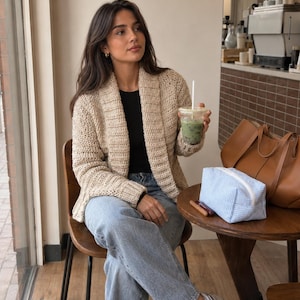 Puede incluir: Un cárdigan de ganchillo beige con cuello chal, una camiseta negra, vaqueros azul claro y zapatillas deportivas grises. Un bolso de cuero marrón y un estuche con cremallera de textura azul claro sobre una mesa redonda de madera.