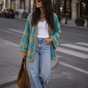 Puede incluir: Un cárdigan de ganchillo con patrón de cuadrados granny en tonos turquesa, naranja y amarillo, usado sobre una camiseta sin mangas blanca y vaqueros azul claro, combinado con un bolso hobo de ante marrón y zapatillas blancas, con gafas redondas.