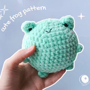 May include: A mint green, crocheted frog plush toy with black eyes and a small smile. The text "cute frog pattern" is written in white script above the frog. The frog is held in a hand against a light blue background.