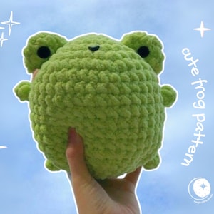 Könnte beinhalten: Eine handgefertigte, hellgrüne Häkelfrosch mit schwarzen Augen und kleinen Gliedmaßen. Der Frosch wird in einer Hand vor einem blauen Himmelshintergrund mit dem Text "cute frog pattern" gehalten. Der Frosch ist ein Plüschtier.