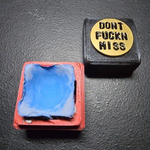 May include: Two small, square objects. One is red with blue material inside. The other is black with a gold-colored circular emblem that reads "DONT FUCKN MISS" in bold, black letters.