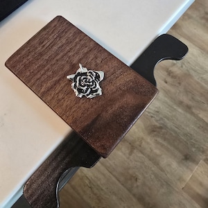 Puede incluir: Un colgante de rosa plateada y negra descansa sobre una superficie de madera marrón oscuro. El colgante está detallado con esmalte negro y cuelga de un pequeño lazo. La superficie de madera es parte de una estructura más grande.