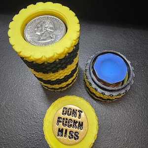 May include: Three coin holders in yellow, black, and grey. One cylindrical holder contains a coin. Another is shorter with a blue top. The third has the text "DONT FUCKN MISS" on a gold-coloured disc.