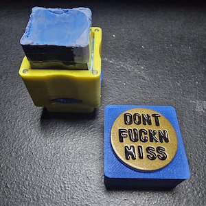 May include: A stacked, yellow and blue cube-shaped object with a blue top. A separate blue square holds a gold-colored disc with the words "DONT FUCKN MISS" in black.