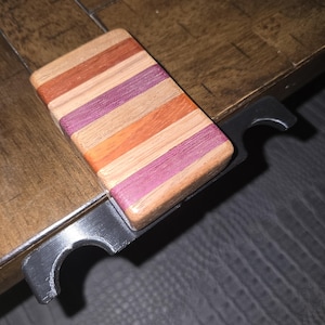 May include: A bottle opener featuring a rectangular handle made of alternating horizontal strips of light tan, reddish-orange, and purple wood. The handle is attached to a black metal opener mechanism with a curved notch for opening bottles.
