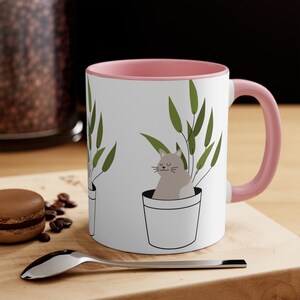 Peut inclure: Un mug en céramique blanche avec un intérieur et une anse rose pâle, orné d'une illustration graphique répétée d'un chat brun clair assis dans un pot de fleurs blanc avec des feuilles vertes s'étendant vers le haut derrière le chat.