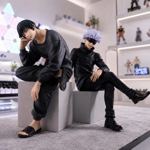 May include: Two anime-style character figures sitting on gray cubes. One figure has short black hair, wearing a long-sleeved black shirt, black pants, and black slides. The other figure has spiky light purple hair, a black blindfold, a black high-neck shirt, black pants, and black boots.