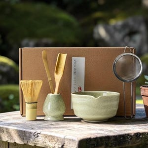 Peut inclure: Un service à thé japonais disposé sur une table en bois. L'ensemble comprend un fouet, une cuillère et une spatule en bambou, un bol en céramique, une passoire en métal et une petite plante succulente en pot. Une boîte en carton est à l'arrière-plan.
