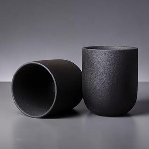 Peut inclure: Deux tasses en céramique noire mate. L'une est droite, l'autre est sur le côté. Les tasses ont une forme cylindrique arrondie avec une surface texturée. Le fond est gris uni.