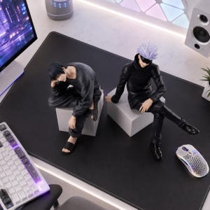 May include: Two anime-style character figures sitting on white cubes. One figure has short black hair, wearing a dark gray long-sleeve shirt, dark pants, and black slides. The other figure has spiky light purple hair, a black blindfold, a black high-neck jacket, black pants, and black shoes.
