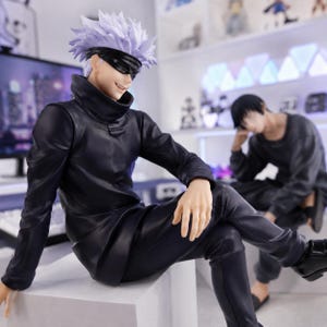 May include: A collectible anime figure of a character with spiky white hair and a black blindfold over their eyes. The figure wears a high-collared black jacket and matching black trousers, sitting in a relaxed pose with one leg crossed over the other.
