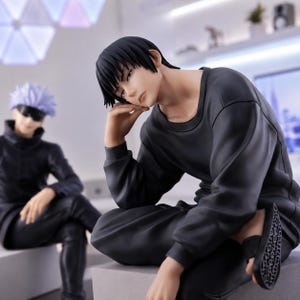 May include: A detailed anime-style figurine of a character with short black hair, wearing a long-sleeved black shirt and black pants, resting their chin on one hand. In the background, a second figurine features a character with spiky light hair.