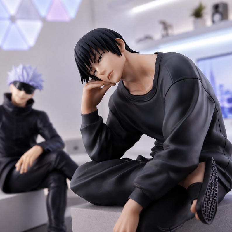 May include: A detailed anime-style figurine of a character with short black hair, wearing a long-sleeved black shirt and black pants, resting their chin on one hand. In the background, a second figurine features a character with spiky light hair.