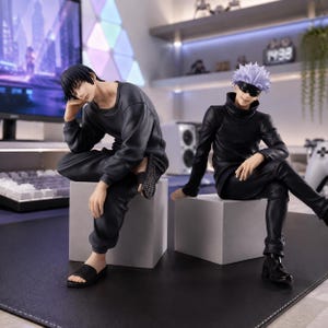 May include: Two anime character figures sitting on gray cubes. The left figure has short black hair, wearing a dark gray long-sleeve shirt, black pants, and black slides. The right figure has spiky light purple hair, a black blindfold, a black high-neck shirt, black pants, and black shoes.