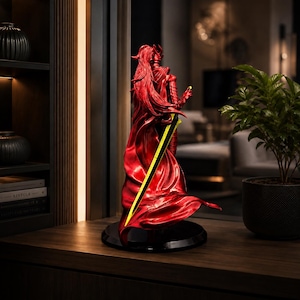 May include: A metallic red figurine of a warrior with long flowing hair and armor, standing on a circular black base. The figure holds a long, slender sword with a bright yellow blade that extends diagonally across the back of the sculpture.