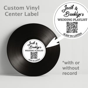 Puede incluir: Un disco de vinilo negro con una etiqueta central blanca que dice "Jacob & Brooklyn's WEDDING PLAYLIST" y un código QR. El texto "SCAN TO LISTEN" está debajo. Las palabras "Custom Vinyl Center Label" están en la parte superior.