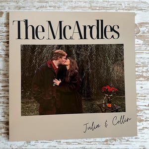 Könnte beinhalten: Ein gerahmtes Foto mit dem Text "The McArdles" oben und "Julia & Collin" unten. Das Foto im Inneren des Rahmens zeigt ein sich küssendes Paar vor einer Trauerweide. Im Hintergrund ein kleiner Tisch mit Blumen und einer Flasche.