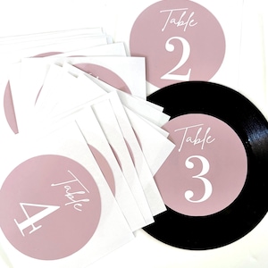 May include: A stack of square white cards featuring dusty rose circular centers with the word Table and a large numeral printed in white. One card is placed on a black circular record-style base, displaying the text Table 3 in white.
